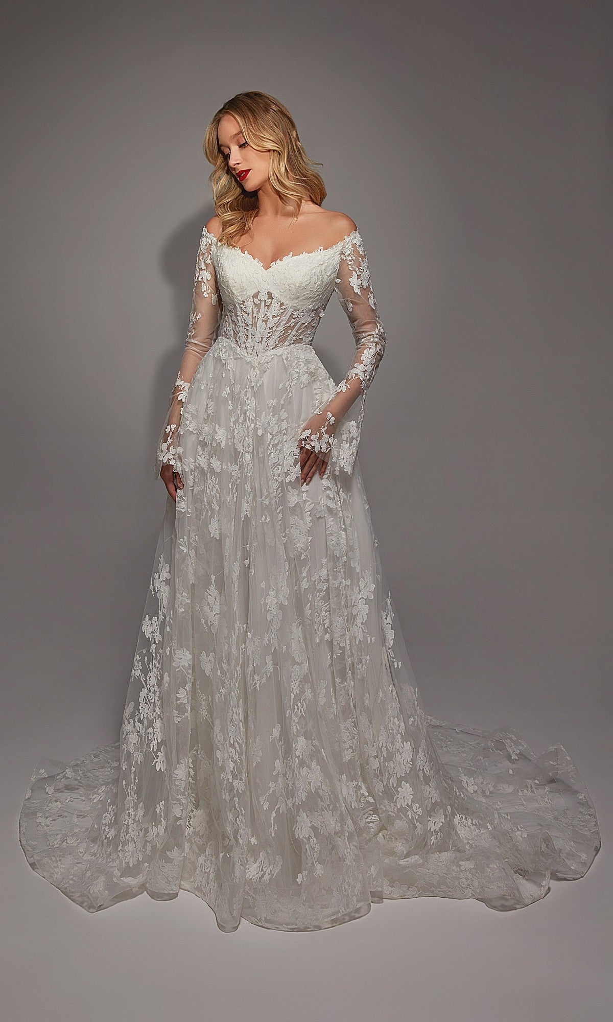 A woman models the Alyce 7150, a long ivory/sand dress with off-the-shoulder long sleeves, white lace, ruffle details on the fitted bodice, and a flowing skirt against a plain background.